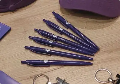 Set de bolígrafos promocionales morados personalizados con logo impreso por Bresspop, exhibidos junto a otros artículos de material POP como gorras y llaveros en una superficie de madera.